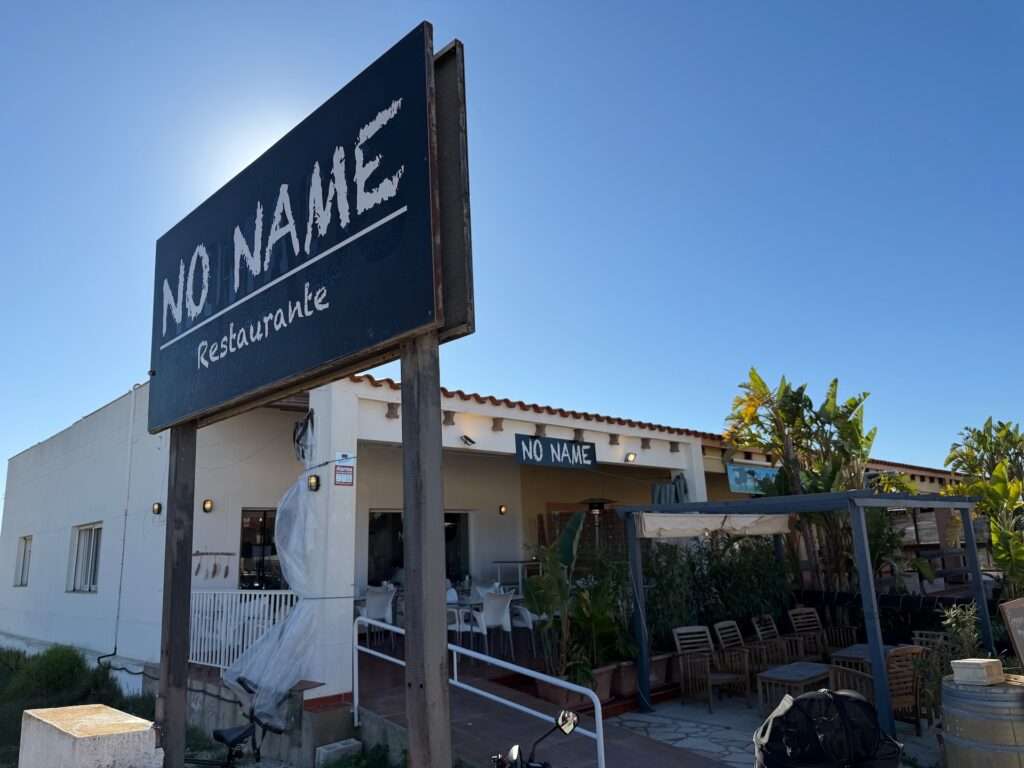 Algarve 2025-26 – 11 Ein großes Schild mit der Aufschrift "NO NAME Restaurante" steht vor einem weißen Gebäude mit einem Sitzbereich im Freien, der von Markisen beschattet wird und von Pflanzen umgeben ist, unter einem klaren blauen Himmel.