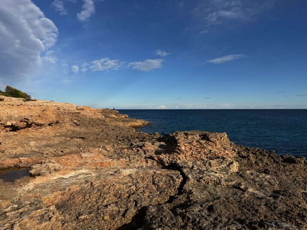 Algarve 2025-26 – 16 Felsige Küstenlinie mit braunen Felsen im Vordergrund, klares blaues Meer in der Mitte und ein teilweise bewölkter blauer Himmel darüber. Das Sonnenlicht wirft Schatten auf die Felsen. Auf der linken Seite ist spärliches Grün zu sehen.