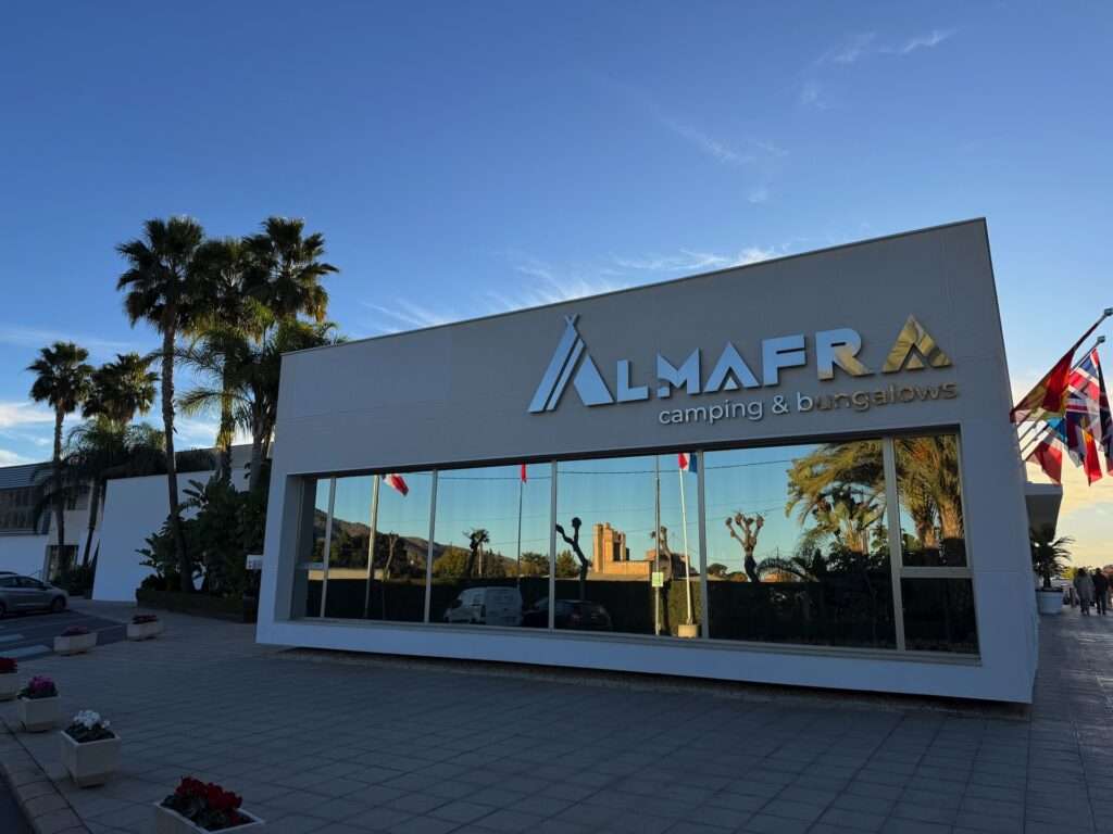 Algarve 2025-26 – 17 Modernes weißes Gebäude mit großen Glasfenstern, in denen sich Palmen und Fahrzeuge spiegeln, mit dem Schild "Almafra camping & bungalows". Auf der rechten Seite sind mehrere Fahnen zu sehen, und Topfpflanzen säumen den Gehweg.