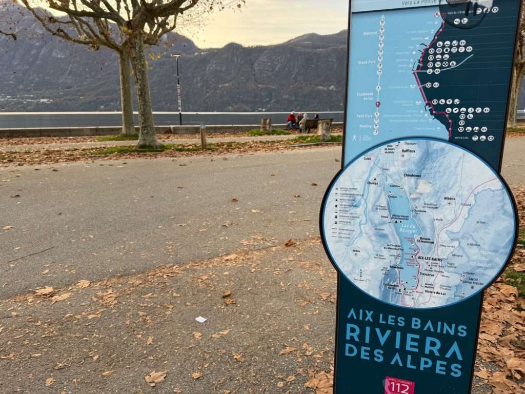 Algarve 2025-26 – 2 Ein Schild "Aix Les Bains Riviera des Alpes" mit einer detaillierten Karte steht neben einem von Bäumen gesäumten Uferweg. Im Hintergrund sind Berge und ein ruhiger See unter einem bewölkten Himmel zu sehen.