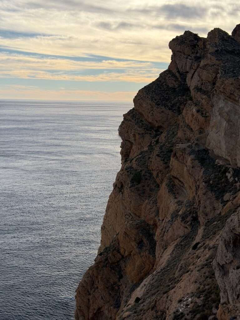 Algarve 2025-26 – 21 Eine zerklüftete Felsklippe erhebt sich steil über dem ruhigen, weiten Meer unter einem teilweise bewölkten Himmel bei Sonnenuntergang.