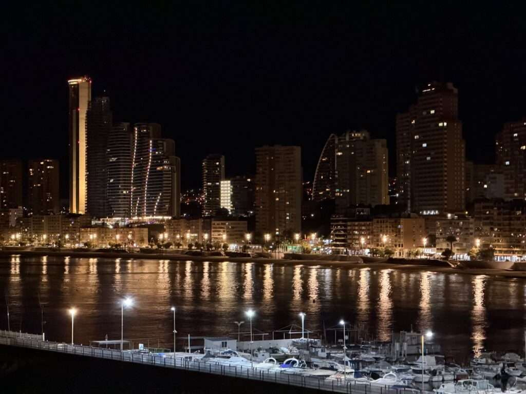 Algarve 2025-26 – 23 Eine nächtliche Stadtlandschaft mit hohen, beleuchteten Gebäuden und einem Riesenrad, wobei sich die Lichter auf dem Wasser spiegeln und die Boote im Vordergrund angedockt sind.