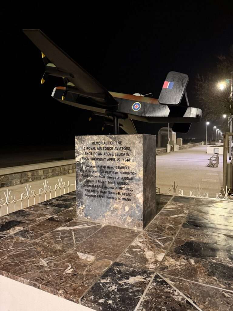 Algarve 2025-26 – 6 Nachtansicht eines steinernen Denkmals mit einem Modellflugzeug aus der Zeit des Zweiten Weltkriegs auf der Spitze zu Ehren der Flieger der Royal Air Force. Der eingravierte Text listet Namen und ein Datum auf. Im Hintergrund sind Straßenlaternen, Bänke und ein Strand zu sehen.