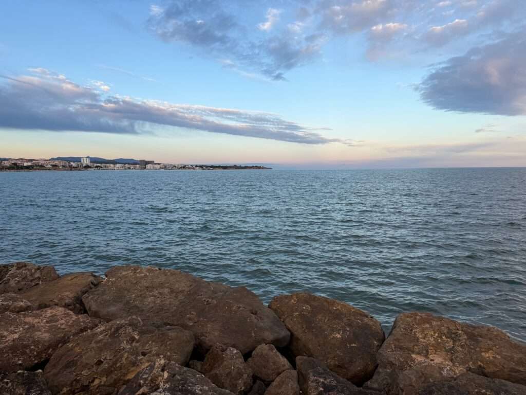 Algarve 2025-26 – 8 Große Felsen im Vordergrund mit ruhigem, blauem Meer, das sich bis zum Horizont erstreckt; eine entfernte Küstenlinie mit Gebäuden unter einem wolkenverhangenen Himmel bei Sonnenuntergang oder in der Abenddämmerung.