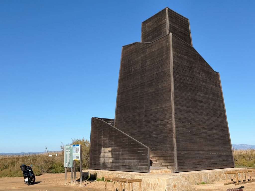 Algarve 2025-26 – 9 Ein großer, moderner hölzerner Aussichtsturm steht auf einer unbefestigten Fläche mit strahlend blauem Himmel. Ein Fahrrad mit Gepäcktaschen ist in der Nähe einer Informationstafel und von Bäumen im Hintergrund abgestellt.
