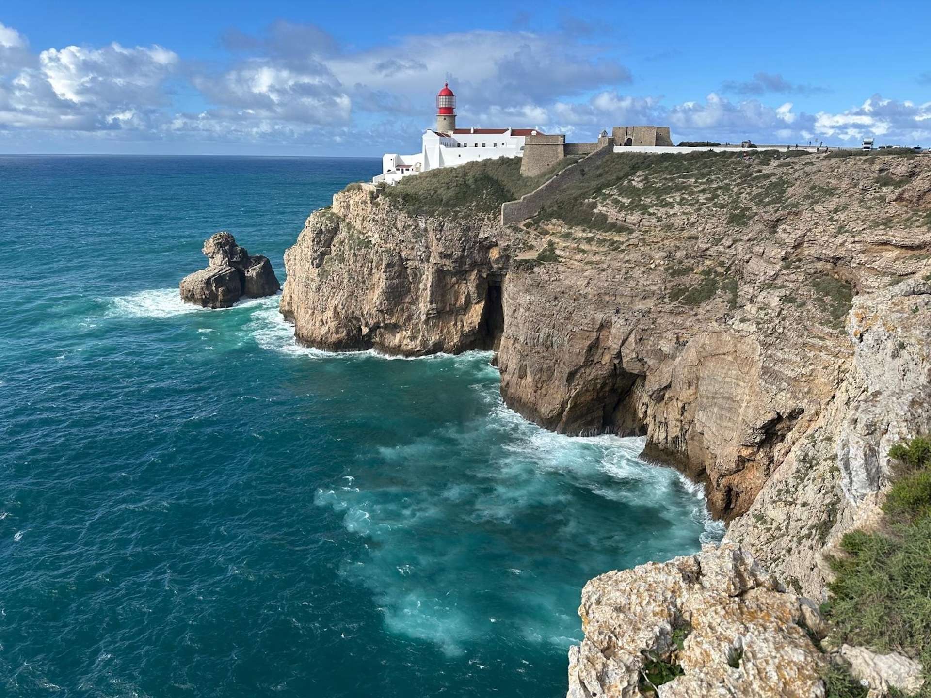 Ein weißer Leuchtturm mit roter Kuppel steht auf einer zerklüfteten Klippe und überblickt das türkisfarbene Wasser des Ozeans unter einem teilweise bewölkten blauen Himmel. Die Wellen schlagen gegen die felsige Küste.