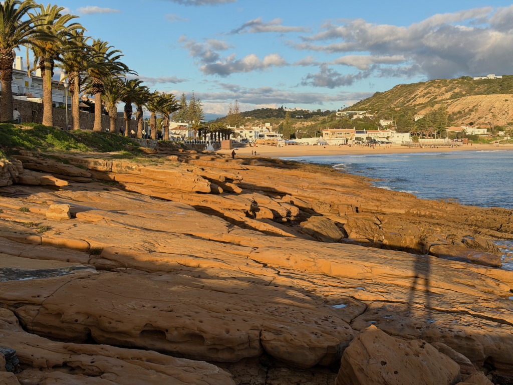 Goldene Sandsteinfelsen erstrecken sich entlang eines sonnenbeschienenen K&uuml;stenstrandes mit Palmen an der Promenade, wei&szlig;en Geb&auml;uden und sandigen Klippen im Hintergrund unter einem teilweise bew&ouml;lkten Himmel.