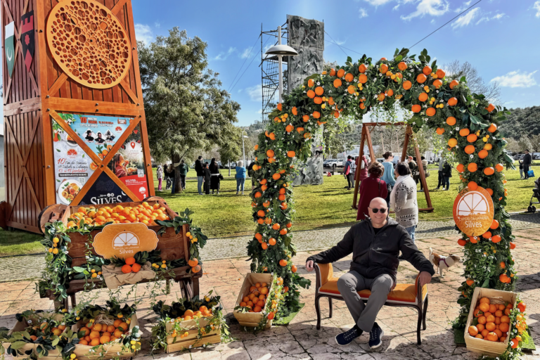 Ein Mann sitzt auf einer Bank unter einem mit Orangen und grünen Blättern geschmückten Bogen bei einem Fest im Freien. In der Nähe stehen mit Orangen gefüllte Kisten. Im Hintergrund sind an einem sonnigen Tag Menschen versammelt.