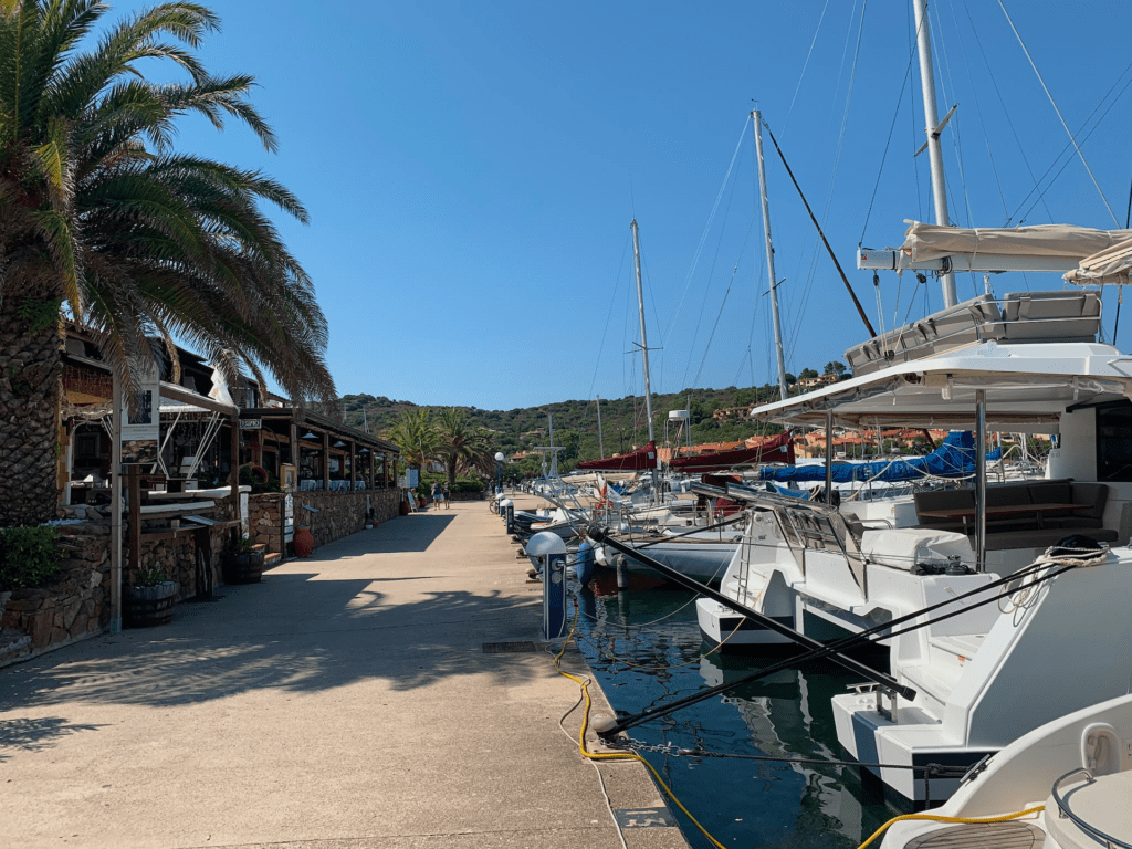 072c3a5e-c59d-480d-a055-0f44f7280759 Eine sonnige Szene in einem Yachthafen mit am Wasser vor Anker liegenden Booten, einer Palme und einem Steinweg neben einem rustikalen Restaurant am Wasser. Im Hintergrund sind blauer Himmel und grüne Hügel zu sehen.