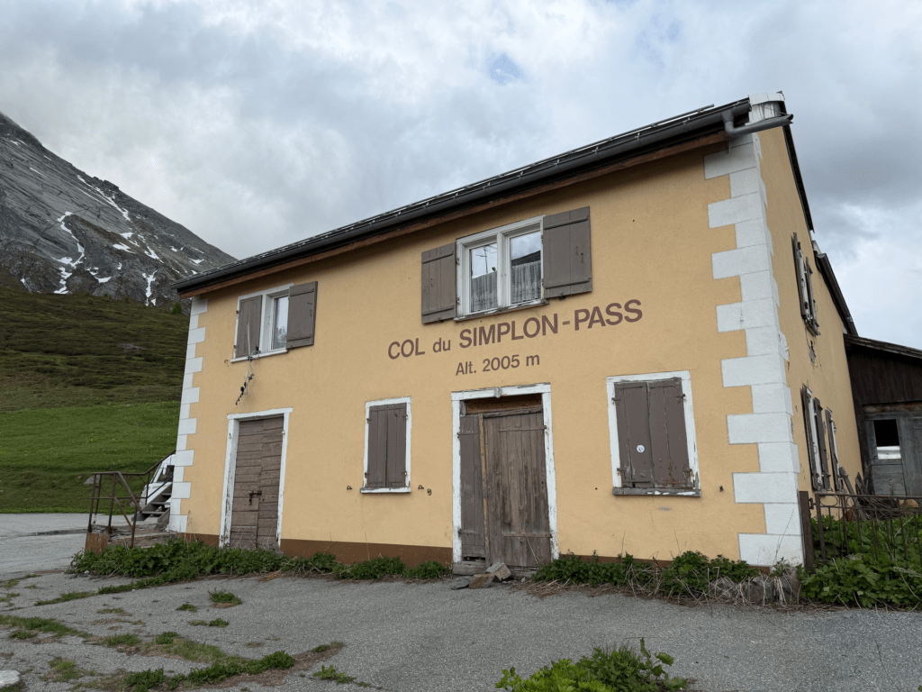 IMG_5241 An der Wand eines gelben Gebäudes mit braunen Fensterläden steht „COL du SIMPLON-PASS, Höhe 2005 m“. Das Gebäude steht vor einem grasbewachsenen Hügel und einem bewölkten Himmel, wobei links ein Berg teilweise zu sehen ist.