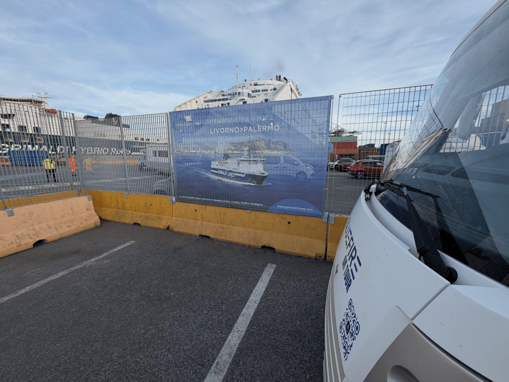 IMG_5271 Hinter einem Zaun, an dem ein großes Schild für die Fährverbindung Livorno–Palermo angebracht ist, liegt eine Fähre vor Anker. Rechts davon steht ein weißes Fahrzeug, und links stehen zwei Arbeiter in gelben Westen in der Nähe von Absperrungen.