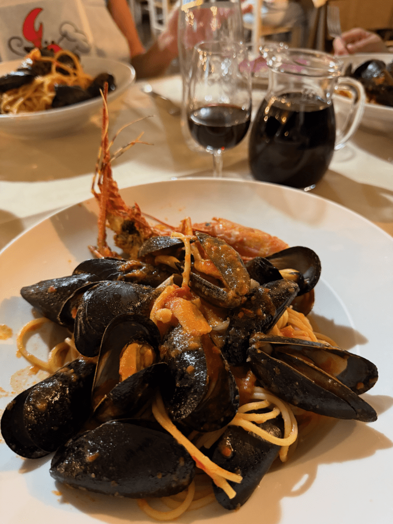 IMG_5361 Auf einem Tisch in einem Restaurant steht ein Teller mit Meeresfrüchte-Pasta mit Muscheln und Garnelen, dazu ein Glas und ein Krug Rotwein. Im Hintergrund ist ein weiteres Pastagericht zu sehen.